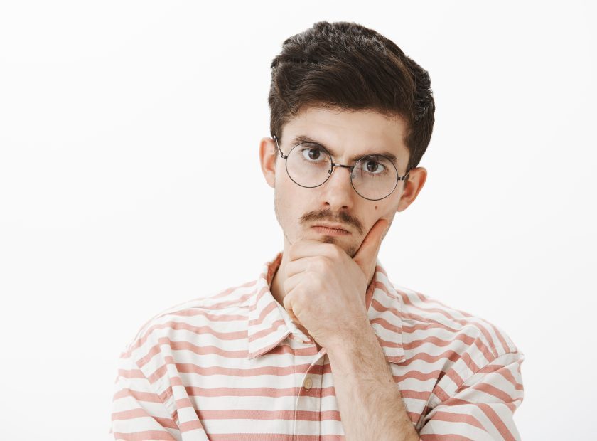 Indoor shot of serious focused angry elder brother with moustache in trendy glasses, holding hand on chin and staring at camera with displeased annoyed expression, solving hard problem over gray wall