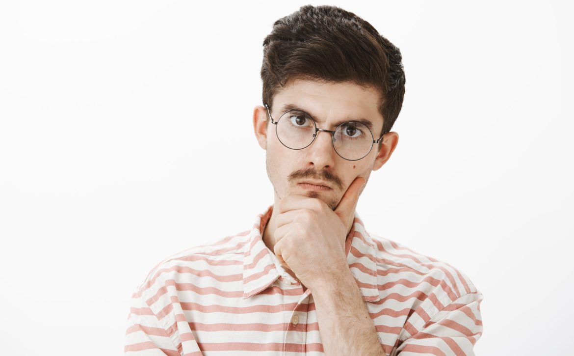 Indoor shot of serious focused angry elder brother with moustache in trendy glasses, holding hand on chin and staring at camera with displeased annoyed expression, solving hard problem over gray wall