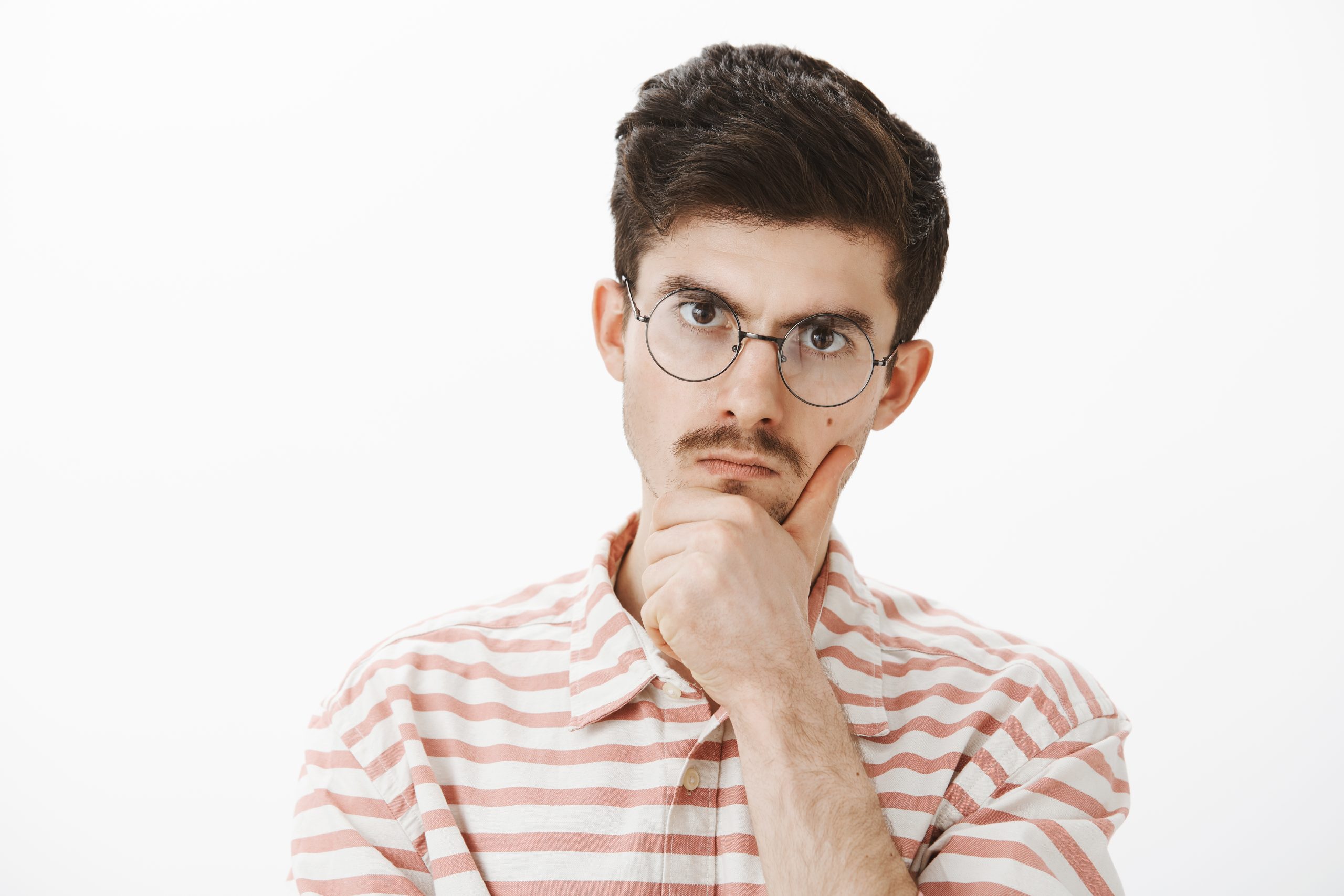 Indoor shot of serious focused angry elder brother with moustache in trendy glasses, holding hand on chin and staring at camera with displeased annoyed expression, solving hard problem over gray wall